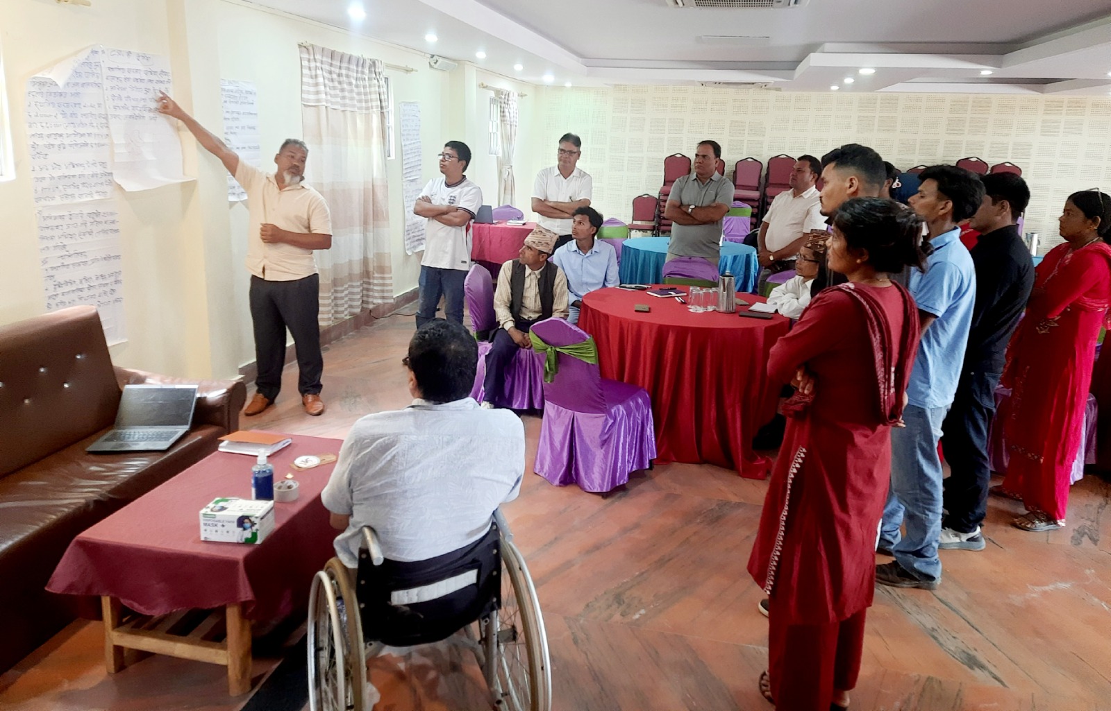 A man presenting in front of a chart paper posted on a wall to a group of workshop participants sitting and standing in a meeting room, including a person in a wheelchair in the foreground.